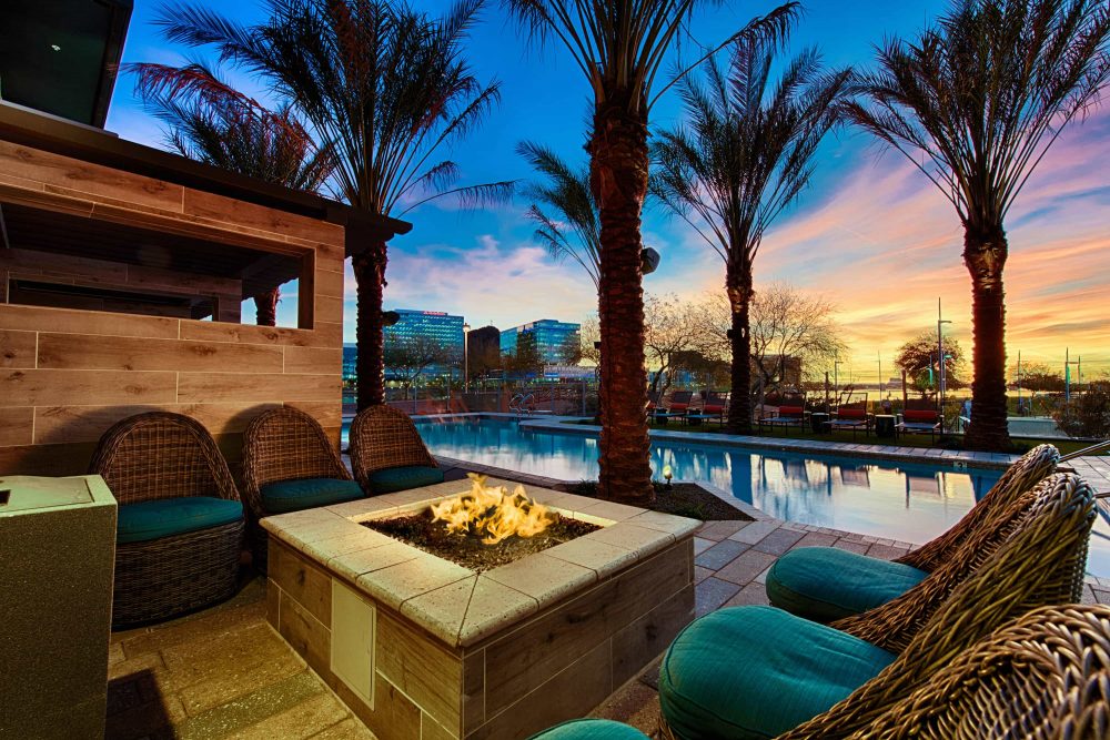Tempe, AZ Luxury Apartments - Vela At Tempe Town Lake - Poolside Lounge Area With Raised Firepit, Outdoor Seating, Palm Trees, And City View